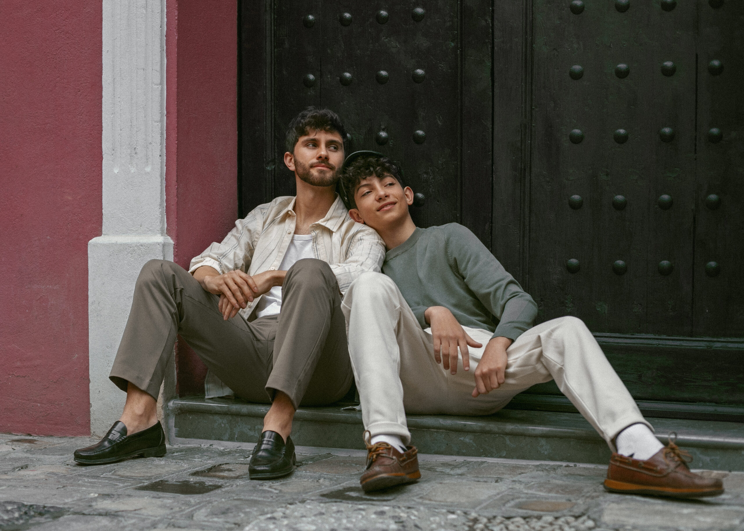 Two men sit closely on a stone step, one resting his head on the other's shoulder, sharing a calm, affectionate moment outdoors.