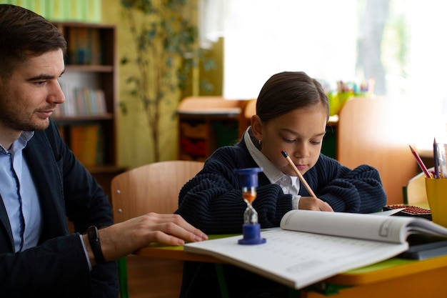 Teacher assisting a young student who is focused on writing in a notebook at a classroom desk.