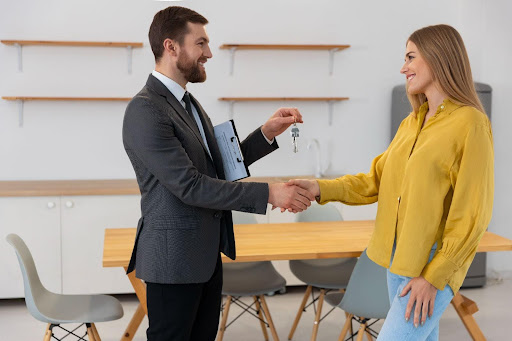A real estate agent handing keys to a woman while shaking her hand in a modern, empty home.