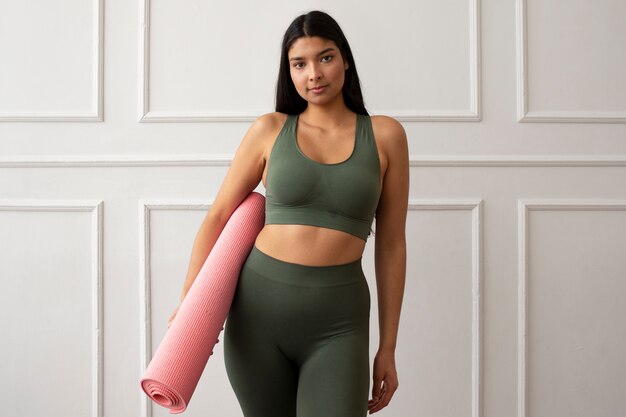 Person wearing a green sports bra and leggings, holding a rolled yoga mat indoors.