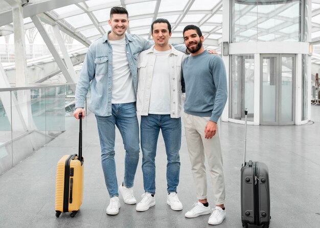 Three men standing together with luggage in a modern glass walkway, smiling and wearing casual outfits.