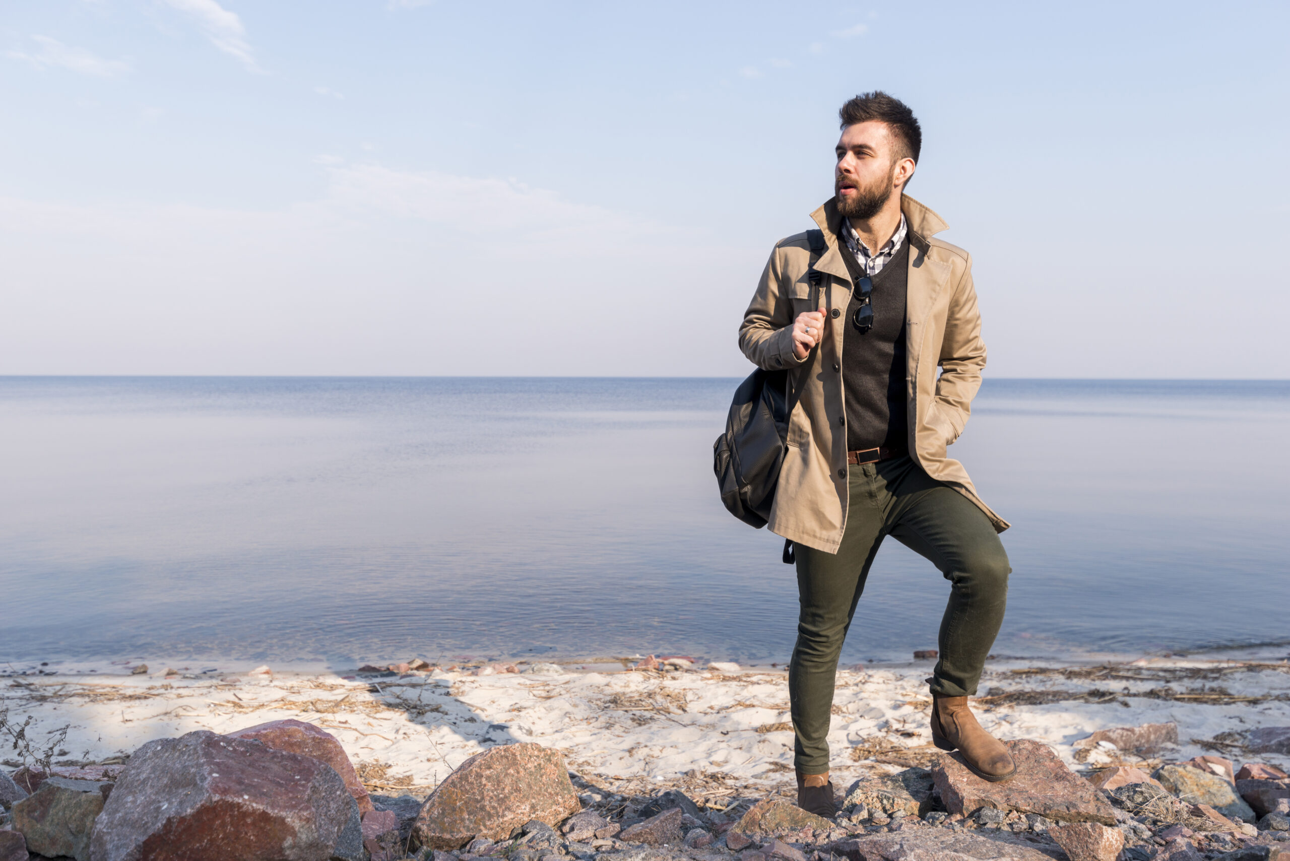 Man wearing a beige coat, dark sweater, and green pants stepping on rocks by a calm sea, carrying a backpack and looking aside.