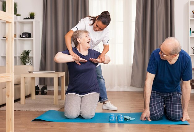 Caregiver helping a senior woman exercise on a yoga mat at home, while an elderly man watches and smiles.