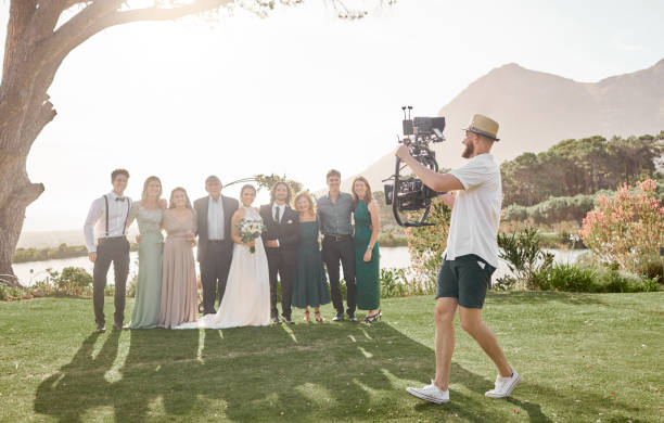 Wedding photographer filming a group of guests and a couple outdoors with mountains and a lake in the background.