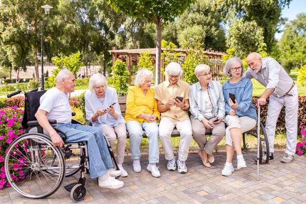 Group of senior friends sitting on a park bench using smartphones and smiling together in a sunny garden.